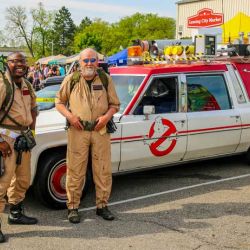 People in Ghostbusters costumes standing next to Ecto-1 replica car at 517 Day event